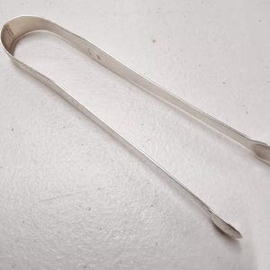 May include: Silver sugar tongs with a rounded, slightly curved design. The tongs are made of polished silver and have a smooth, shiny finish.