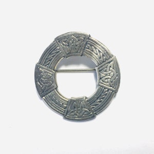 May include: A silver-toned circular brooch with a Celtic knot design. The brooch features a pin on the back for secure fastening. The design includes intricate knotwork patterns within a circular frame, a classic piece of jewelry.
