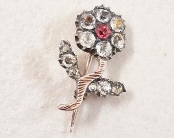 May include: A silver and gold flower brooch with a red center stone and clear stones surrounding it. The brooch has a stem with leaves.