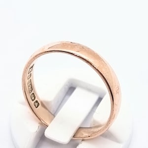 May include: A rose gold ring with a smooth, polished surface. The ring is displayed on a white stand. The inside of the ring has several stamped markings. This ring is a piece of jewelry.