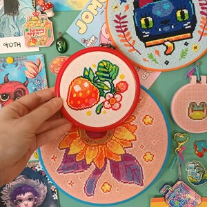 May include: A collection of colorful cross-stitch embroidery hoops. The designs include a strawberry, a sunflower, a cat, and a frog. Other items include a keychain with a mushroom and a carton of honey apple tea.