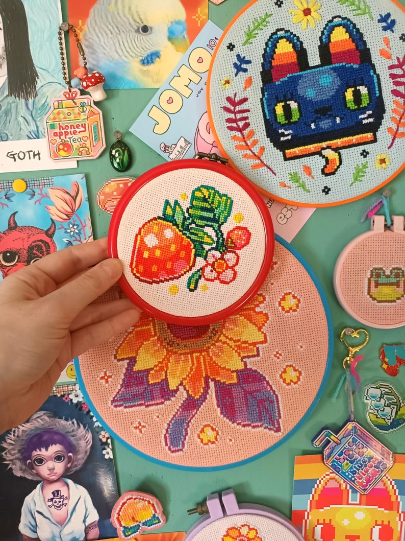 May include: A collection of colorful cross-stitch embroidery hoops. The designs include a strawberry, a sunflower, a cat, and a frog. Other items include a keychain with a mushroom and a carton of honey apple tea.