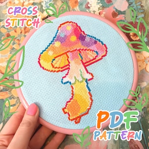 Op de afbeelding: Een kruissteek paddestoelontwerp in een roze borduurring. De paddestoel heeft een kleurrijk pixelontwerp met gele, oranje, roze, paarse en blauwe tinten. De tekst "CROSS STITCH" en "PDF PATTERN" zijn zichtbaar.