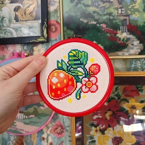 May include: A cross-stitch embroidery of a strawberry with green leaves and pink flowers, set in a red hoop. The needlework is on a cream-colored fabric. The background includes framed artwork and floral wallpaper.