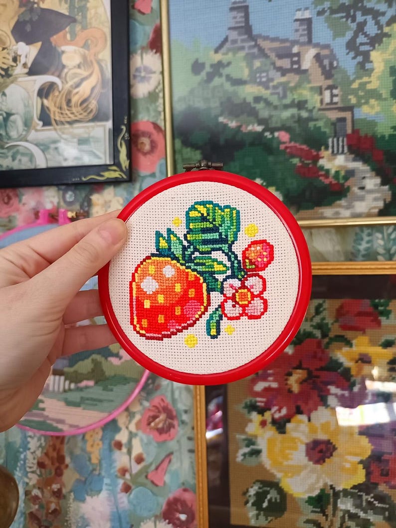 May include: A cross-stitch embroidery of a strawberry with green leaves and pink flowers, set in a red hoop. The needlework is on a cream-colored fabric. The background includes framed artwork and floral wallpaper.