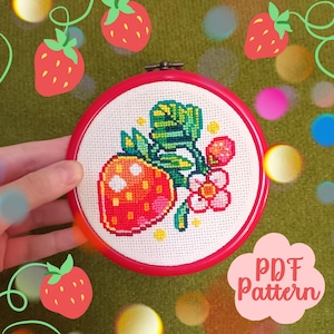 May include: A cross-stitch embroidery hoop featuring a vibrant strawberry design. The red hoop frames a white fabric with a detailed cross-stitch of a red strawberry, green leaves, and a pink flower. The text "PDF Pattern" is visible.