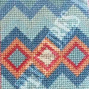 Op de afbeelding: Een kruissteekpatroon met een geometrisch ontwerp met blauwe, oranje en rode zigzaggen en diamanten. Het patroon is omlijst met een witte rand en een rode en oranje rand.