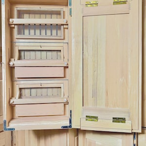 May include: A wooden cabinet with a hinged door that opens to reveal three shelves with mesh screens. The cabinet is made of light wood and has brass hinges.