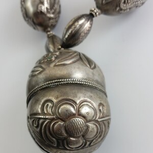 May include: A silver necklace with a large, ornate pendant. The pendant is shaped like an egg and is decorated with intricate floral designs. The necklace is made of silver beads and has a silver clasp.