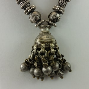 May include: A silver necklace featuring a bell-shaped pendant with numerous small bells hanging from it. The necklace is crafted from silver beads and displays a traditional design.