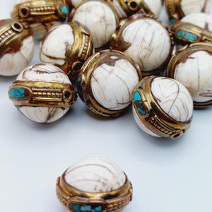 Puede incluir: Una colección de cuentas de hueso blanco con detalles dorados e incrustaciones de turquesa. Las cuentas son redondas y tienen una superficie texturizada.