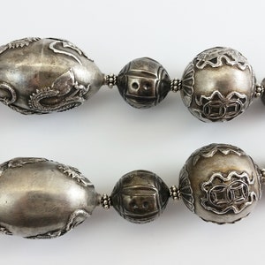 May include: A silver necklace with large, intricately carved beads. The beads are oval and round in shape and have a textured surface.