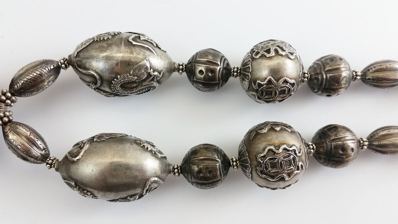 May include: A silver necklace with large, intricately carved beads. The beads are oval and round in shape and have a textured surface.