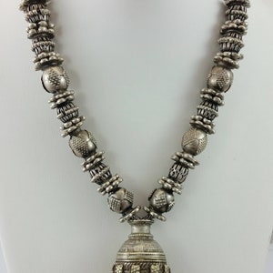 May include: A silver necklace with a pendant featuring a bell-shaped charm with multiple dangling beads. The necklace is made of round beads with intricate designs.