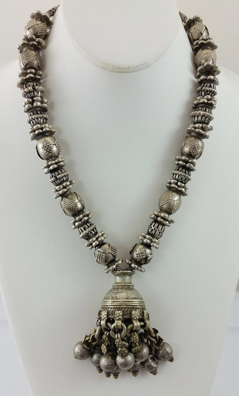 May include: A silver necklace with a pendant featuring a bell-shaped charm with multiple dangling beads. The necklace is made of round beads with intricate designs.