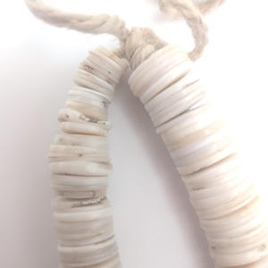 May include: Close-up of a necklace made of white, flat, circular shell beads strung on a beige rope. The beads are stacked together, creating a curved shape. The rope is knotted at the top.