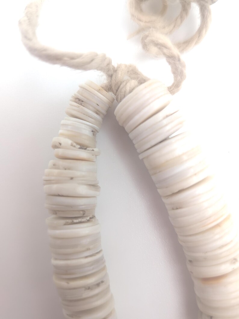 May include: Close-up of a necklace made of white, flat, circular shell beads strung on a beige rope. The beads are stacked together, creating a curved shape. The rope is knotted at the top.