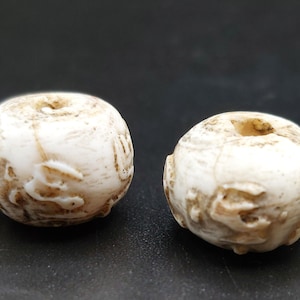 Puede incluir: Dos cuentas redondas blancas con tallas intrincadas. Las cuentas tienen un agujero en el centro para enhebrar.