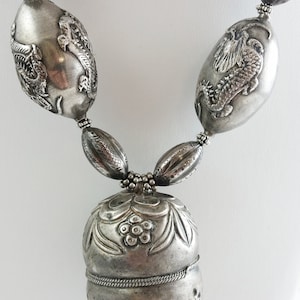 May include: A silver necklace with a large, ornate pendant. The pendant is round and has a detailed floral design. The necklace is made of silver beads and has a silver chain.
