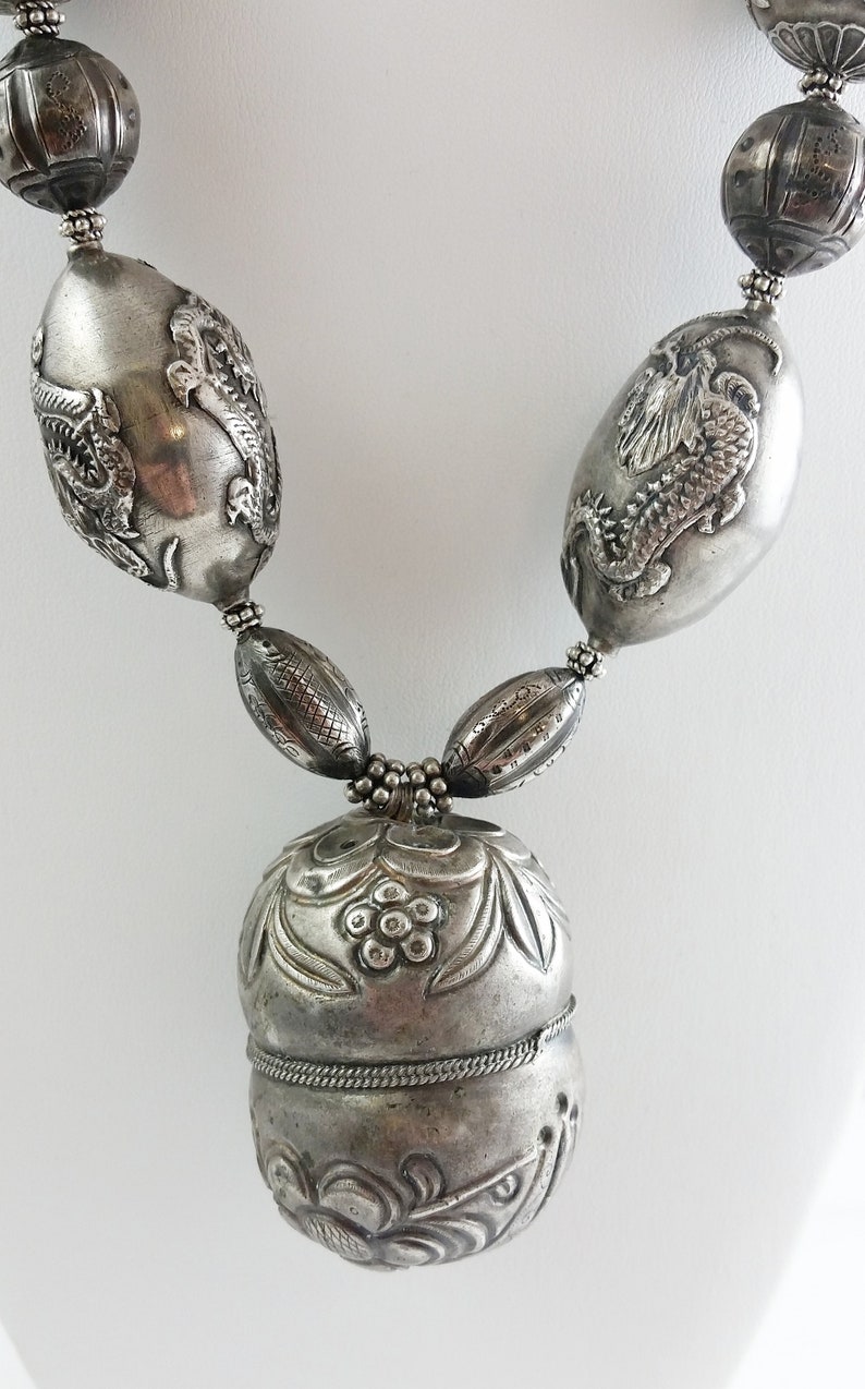 May include: A silver necklace with a large, ornate pendant. The pendant is round and has a detailed floral design. The necklace is made of silver beads and has a silver chain.