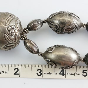 May include: A silver necklace with large, intricately carved beads. The beads are oval-shaped and feature floral and geometric designs. The necklace is approximately 6 inches long.