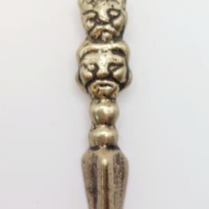 May include: A small, antique-style metal pendant with a loop at the top. The pendant features two carved faces stacked on top of each other, with a pointed, dagger-like bottom. The metal has a tarnished, aged appearance.