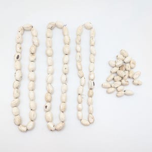 Puede incluir: Tres hilos de cuentas oblongas blanquecinas, junto a un montón de cuentas sueltas, todo sobre un fondo blanco. Las cuentas tienen un aspecto natural y orgánico, con variaciones de color y textura, lo que sugiere un material artesanal o natural.