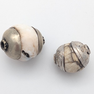 Puede incluir: Dos cuentas antiguas. Una es una cuenta grande, ovalada, con un centro blanco y beige y tapas de metal plateado. La otra es una cuenta más pequeña, en forma de barril, con un diseño similar y detalles metálicos. Ambas cuentas tienen detalles intrincados.