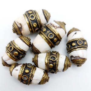 Puede incluir: Seis cuentas de hueso blanco con detalles en latón. Las cuentas son de forma ovalada y tienen un patrón de rayas blancas y marrones. Los detalles en latón son en forma de bandas y círculos.