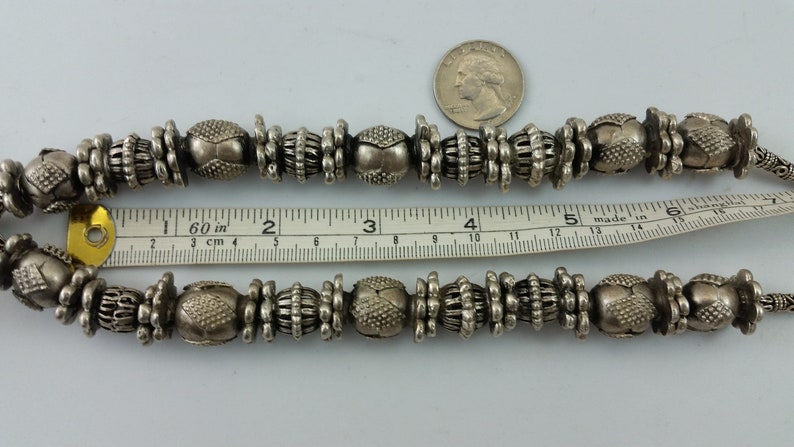 May include: A silver necklace with large, ornate beads. The necklace is approximately 15 inches long and is made of metal beads with a textured surface. The beads are arranged in a repeating pattern of three beads.