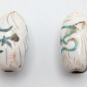 Puede incluir: Dos cuentas ovaladas blancas con detalles turquesa y dorados. Las cuentas tienen un diseño tallado y probablemente son para hacer joyas o manualidades. Las cuentas tienen un aspecto ligeramente envejecido.