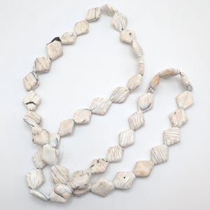 Puede incluir: Un collar con cuentas cuadradas de color crema, con vetas grises oscuras. Las cuentas están ensartadas, creando un diseño largo y fluido. El collar se muestra sobre un fondo blanco, resaltando la textura natural de la piedra.