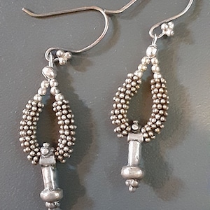 May include: A pair of silver earrings with a beaded design and a teardrop shape. The earrings have a silver hook closure.
