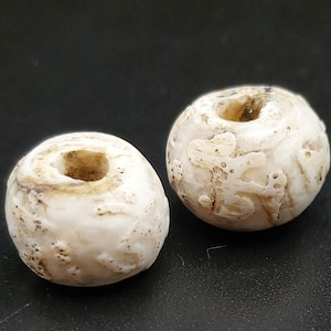 Puede incluir: Dos cuentas de cerámica blanca con un agujero en el centro. Las cuentas tienen una superficie texturizada y parecen ser antiguas.