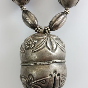 May include: A silver necklace with a large, ornate pendant. The pendant is shaped like a gourd and is decorated with floral and leaf motifs. The necklace is made up of smaller, round beads.