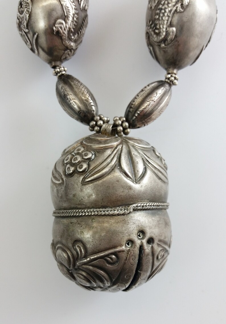 May include: A silver necklace with a large, ornate pendant. The pendant is shaped like a gourd and is decorated with floral and leaf motifs. The necklace is made up of smaller, round beads.