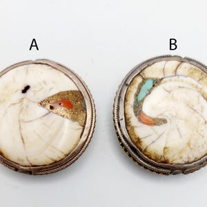 Puede incluir: Dos botones redondos de hueso blanco, con borde plateado, con incrustaciones de turquesa y coral. Los botones tienen un diámetro de aproximadamente 2,5 cm.