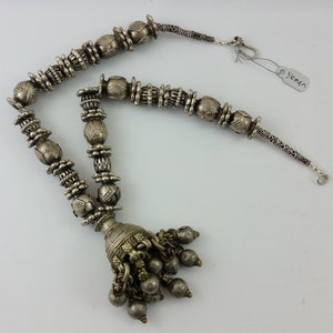 May include: A silver necklace with a tassel pendant and intricate beadwork. The necklace is made of small, round beads with a textured surface. The tassel is made of several small, round beads that are attached to a larger, round bead. The necklace is labelled "Yemen".