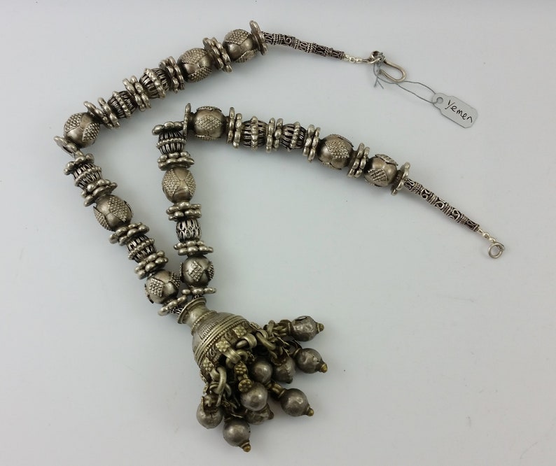 May include: A silver necklace with a tassel pendant and intricate beadwork. The necklace is made of small, round beads with a textured surface. The tassel is made of several small, round beads that are attached to a larger, round bead. The necklace is labelled "Yemen".
