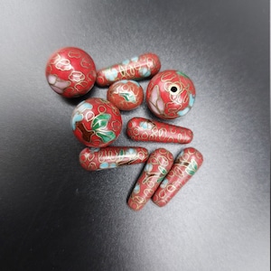 May include: A collection of red, white, and green cloisonné beads with floral designs. The beads are round and oval shaped.