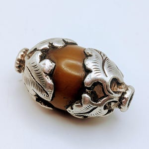 May include: An antique decorative object with a brown, oval centre and silver-coloured metal detailing. The metalwork has leaf-like patterns. The object has a small, textured knob on each end.