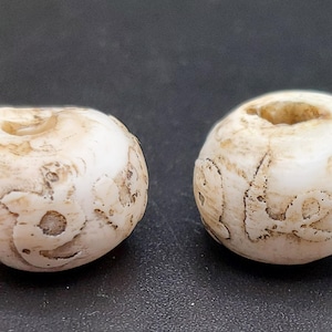 Puede incluir: Dos cuentas redondas blancas con diseños tallados. Las cuentas tienen un agujero en el centro para enhebrar.