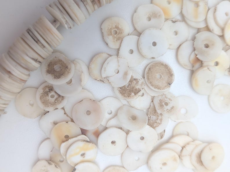 May include: A collection of off-white, disc-shaped beads with a central hole, some showing natural markings. A strand of similar beads is visible on the left. These beads are suitable for jewelry making or craft projects.