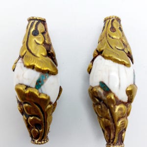 Puede incluir: Dos cuentas doradas y blancas adornadas con intrincados diseños florales. Las cuentas están hechas de piedra blanca con detalles dorados y detalles turquesa.