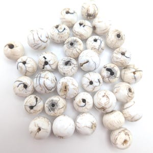 Puede incluir: Un conjunto de cuentas redondas de color blanco roto con vetas marrón oscuro. Las cuentas están dispersas, mostrando sus patrones y texturas únicos. Cada cuenta tiene un agujero para ensartar, perfecto para hacer joyas o manualidades.