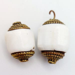 Puede incluir: Dos cuentas de cerámica blanca con detalles en metal dorado. Las cuentas son redondas y tienen un acabado liso y pulido.