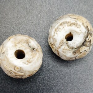 Puede incluir: Dos cuentas redondas de cerámica blanca con un agujero en el centro. Las cuentas tienen una superficie texturizada y están ligeramente desgastadas.