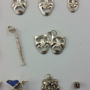 May include: A collection of silver charm pendants featuring various designs, including a clarinet, a piano, a letter 'U', and several variations of comedy and tragedy masks.