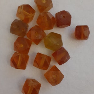 May include: A collection of 13 amber-colored faceted glass beads. The beads are shaped like small cubes with a hole in the center. The beads are scattered on a white surface.