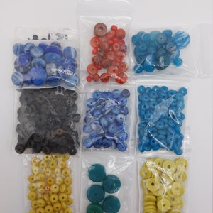 Pode incluir: Conjunto de contas de vidro e madeira coloridas em sacos de plástico individuais. As contas são de várias formas e tamanhos, incluindo redondas, planas e cilíndricas. As cores incluem azul, vermelho, amarelo, verde e preto.